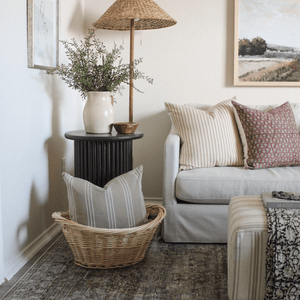 The Darcy pillow is inside a basket in front of a side table with a vase and a lamp. On the sofa are the Leo and Serena pillows
