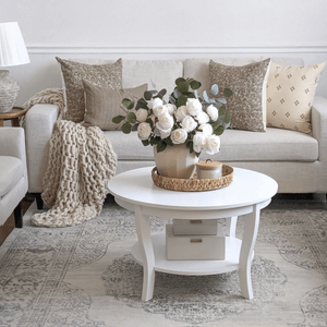 A cozy living room features a beige sofa with woven pillows and a knitted throw. A round white coffee table holds a basket with a vase of white roses and greenery. The space showcases a light, patterned rug and embraces the warmth of Thai cotton in its neutral color palette.