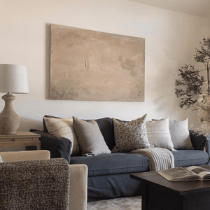 A living room with a dark couch against the far wall. Couch has Rory, Waylon, Forrest, Fitz, and Kinsey pillow. Side table with lamp. Muted abstract painting on back wall.