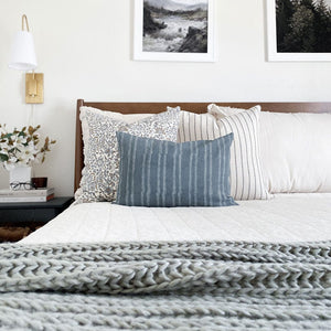 Penelope, Winston, and River lumbar pillow covers from Colin and Finn on cream bed with wood headboard, sage green blanket, gold sconce, and floral arrangement on side table.