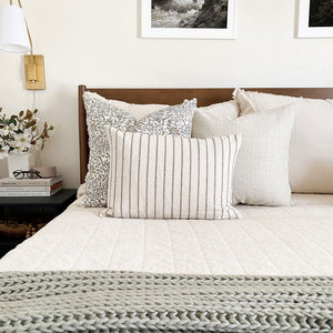 Colin and Finn's Penelope, Selma, and Winston lumbar pillow covers on white bed with wood headboard. Gold sconce and flowers on end table next to bed.