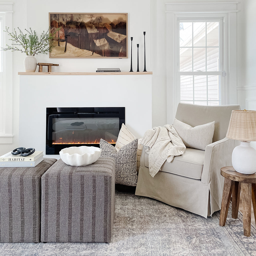 A living room with a fireplace, modern rug, a painting above fireplace, and a light gray chair with Colin and Finn pillows on and around it. Other small decor items are shown as well.