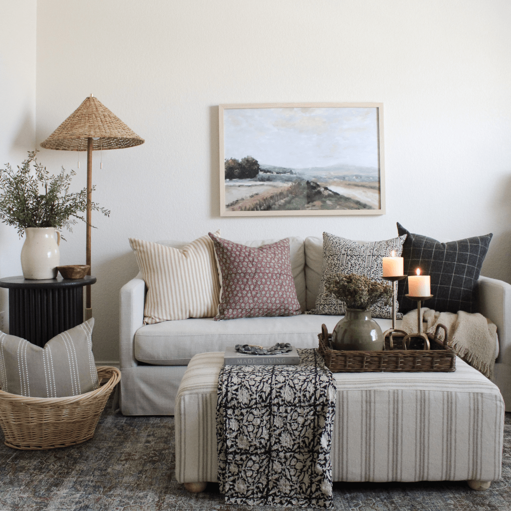 A living room with a sofa centered in the room with four Colin + Finn pillows, Leo, Serena, Madison, and Mavis. A basket in the corner has the Darcy pillow inside. There is an ottoman with a blanket draped across and a basket with candles and a vase with florals.