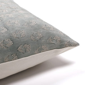 Close-up of a linen pillow cover corner with textured fabric in muted green and beige patterns. The design, reminiscent of traditional printing techniques, features intricate motifs that add elegance to the cushion. The background is a plain, light surface.