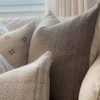 A cozy arrangement of four decorative pillows adorns the couch, including a brown one with a textured design and an Ester Pillow Cover featuring a rust floral motif. Nearby, beige curtains made from natural flax linen frame the scene, adding warmth and elegance to the setting.