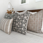 A neatly made bed features a variety of decorative throw pillows with intricate patterns and varying textures. The headboard is a dark wood, complemented by white and neutral-toned pillows. A white wall-mounted lamp and framed artwork are visible in the background.