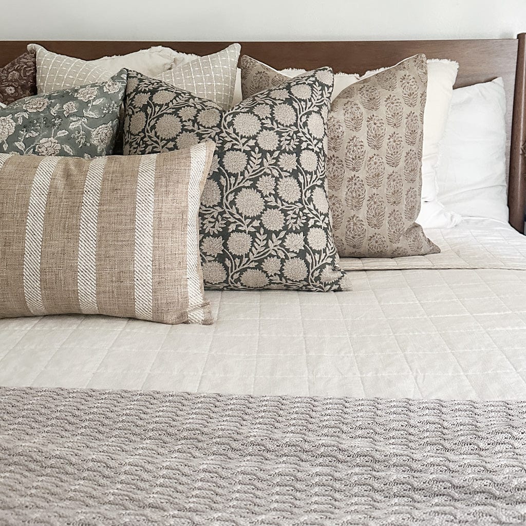 A neatly made bed features a variety of decorative pillows in different patterns and textures. The pillows include designs like florals, stripes, and geometric patterns, all in neutral tones of beige, taupe, and green. The bedspread is white with a light knitted blanket at the foot.