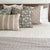 A neatly made bed features a variety of decorative pillows in different patterns and textures. The pillows include designs like florals, stripes, and geometric patterns, all in neutral tones of beige, taupe, and green. The bedspread is white with a light knitted blanket at the foot.