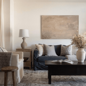 A living room with a dark couch against the far wall. Couch has Rory, Waylon, Forrest, and Kinsey pillow. Coffee table with book and vase. Side table with lamp. Muted abstract painting on back wall. 