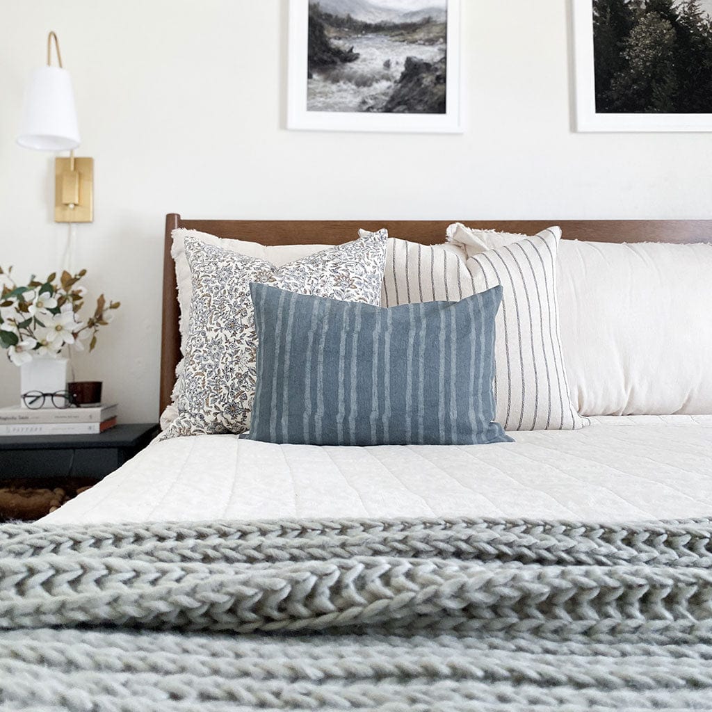 Penelope, Winston, and River lumbar pillow covers from Colin and Finn on cream bed with wood headboard, sage green blanket, gold sconce, and floral arrangement on side table.