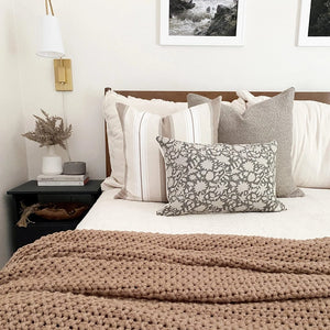 Waylon, Charles, and Sawyer lumbar from Colin and Finn on white bedding with wood headboard. Taupe throw blanket on bed and side table has floral arrangement with gold sconce above.