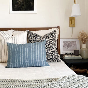 Winston, Edina, and River lumbar pillow cover from Colin and Finn on white bed. Side table has gold sconce, picture frame, and candle.