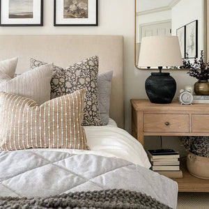 Colin and Finn Bardot in Burlap pillow cover sitting on a king size bed with other throw pillows with a beige headboard and a wood nightstand with black lamp.